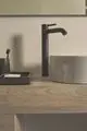 Schwarze Waschbeckenarmatur von Ideal Standard auf Holzoberfläche, neben grauem Waschbecken und schwarzem Organizer mit Utensilien. Schlichte, funktionale Anordnung.